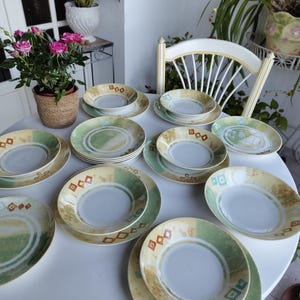 May include: A collection of ceramic plates and bowls with a green, tan, and brown design. The plates are arranged on a white table, with a variety of sizes and shapes. A few bowls are also present. The set is suitable for serving meals.