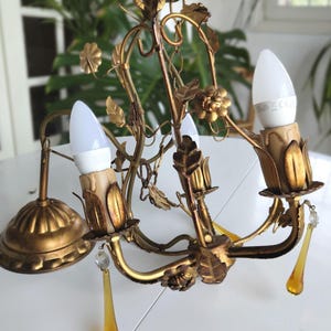 Peut inclure: Un lustre doré avec un design floral. Le lustre comporte trois ampoules et des gouttes de cristal ambré décoratives. Le luminaire est en métal et présente une esthétique vintage.