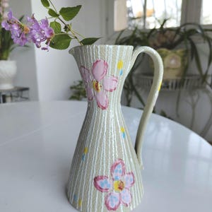 Peut inclure: Un pichet en céramique avec une surface beige clair texturée ornée de rayures verticales blanches et de fleurs roses peintes à la main avec des cœurs jaunes. Le pichet possède une anse incurvée et un bord évasé, contenant une branche de lantana violet.