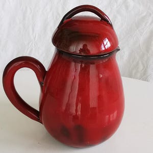 May include: A glossy, red ceramic pitcher with a matching lid and a curved handle. The pitcher has a bulbous body and a dark brown rim. The handle is arched, and the lid has a rounded handle. The pitcher is likely used for serving beverages.
