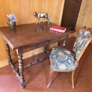 May include: A dark wood desk with a drawer, a brass horse figurine, and a red book. A vase of purple flowers sits on the desk. A patterned chair with a wooden frame is next to the desk. The room has wood paneling and a tiled floor.