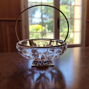 Op de afbeelding: Een heldere glazen mand met een decoratieve goudkleurige metalen voet en handvat. De kom heeft een sterpatroon aan de onderkant en een gouden rand. Het handvat is gemaakt van gedraaid goudkleurig metaal. De mand staat op een bruine houten tafel.