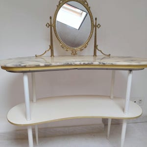 May include: A vintage dressing table with an oval mirror. The table features a marble top with gold trim, a white lower shelf, and white legs. The mirror is framed in gold with an ornate design.