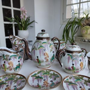 Może przedstawiać: Kompletny, zabytkowy zestaw do herbaty składający się z czajniczka, śmietanki, cukiernicy, filiżanek i spodków. Biały zestaw porcelanowy zdobiony japońskim wzorem z postaciami, glicyniami i złotymi obwódkami.