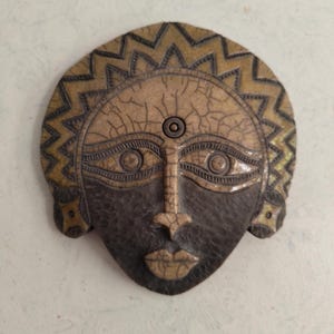 Peut inclure: Un masque décoratif avec un visage. Le masque présente un visage brun foncé avec un nez et des lèvres brun clair. Le masque a une surface craquelée brun clair et un ornement de tête décoratif doré et brun.