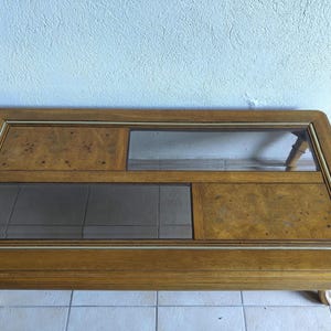 May include: A rectangular coffee table featuring a wooden frame with a light brown finish. The tabletop is composed of alternating sections of burl wood veneer panels and inset glass panels, accented with thin metallic trim along the edges.