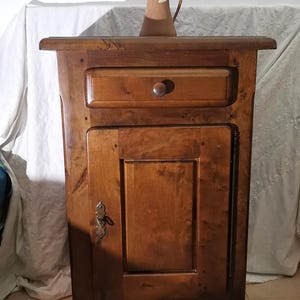Peut inclure: Une table de chevet en bois avec un tiroir et une porte d'armoire. La table de chevet est en bois foncé et possède une petite cruche marron sur le dessus. Le tiroir a un bouton rond en bois. La porte de l'armoire a un loquet décoratif en métal.