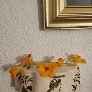 Puede incluir: Un macetero de pared de cerámica blanca con un diseño floral verde y una silueta de pájaro. El macetero está decorado con flores naranjas.