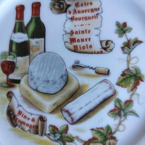 Może przedstawiać: Biały talerz ceramiczny z kolorową ilustracją dwóch butelek wina, kieliszka czerwonego wina, korkociągu i dwóch rodzajów sera. Grafika zwoju zawiera tekst Chinon, Cotes d'Auvergne, Bourgueil, Sainte Maure, Niolo.