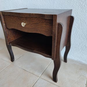 Peut inclure: Une table de chevet en bois brun foncé dotée d'un tiroir unique avec un petit bouton rond de couleur claire. Le meuble présente une surface supérieure plate, une étagère de rangement ouverte sous le tiroir et quatre pieds incurvés.