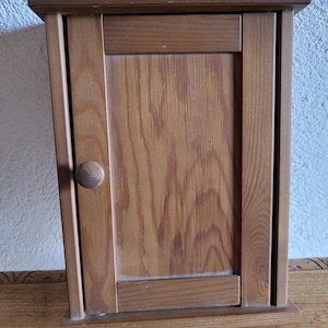 Può includere: Un piccolo mobiletto da parete in legno con un'unica anta e un pomello rotondo in legno. Il mobile è realizzato in legno marrone chiaro con venature visibili e un design rettangolare semplice. Il mobile misura circa 30 cm di altezza.