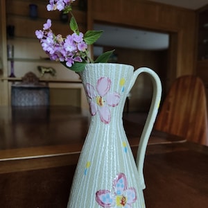 Peut inclure: Un pichet décoratif en céramique vert clair, orné de motifs de fleurs roses et de rayures verticales. Il contient une branche de fleurs violettes avec des feuilles vertes. Le pichet est posé sur une surface en bois brun foncé.