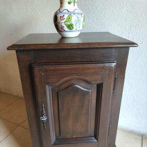 Könnte beinhalten: Ein dunkelbrauner Holzschrank mit einer einzelnen Tür und einem Metallgriff, darauf steht eine cremefarbene Keramikvase mit handgemalten blauen, grünen und kastanienbraunen Blumenmustern und einem blauen Rand an der oberen Öffnung.
