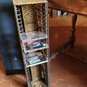 May include: A tall, narrow CD rack with a woven wicker front and sides, and a metal frame. The rack holds several CDs. The rack has a handle at the top and sits on a tiled floor.