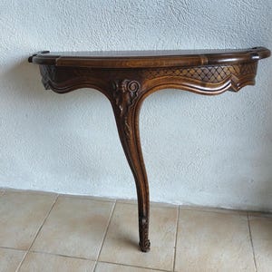 Peut inclure: Une console en bois brun foncé dotée d'un plateau semi-circulaire et d'un pied unique incurvé. La ceinture est ornée d'un motif en treillis sculpté et d'un motif central à volutes. Le grain du bois est visible sur toute la structure.