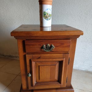 Peut inclure: Une table de chevet en bois au fini brun chaud, dotée d'un tiroir avec une poignée en laiton et d'une porte de placard inférieure avec un petit bouton. Un vase cylindrique en céramique blanche avec un bord en terre cuite est posé dessus.