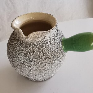 Peut inclure: Petite cruche en céramique texturée avec une poignée verte. La cruche a un extérieur moucheté blanc cassé et gris et un bord beige clair. La poignée est vert brillant. La cruche est probablement utilisée pour servir des liquides.