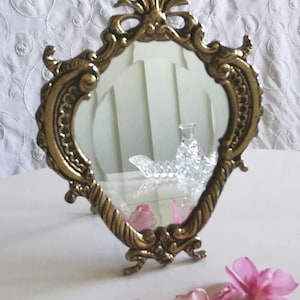 Peut inclure: Un miroir de coiffeuse doré et orné, de forme ovale. Le cadre présente des volutes complexes et un sommet décoratif. Des pétales de fleurs roses sont dispersés près de la base du miroir, ajoutant une touche de couleur.