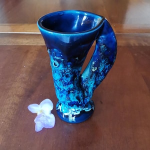 Peut inclure: Un vase en céramique bleu foncé avec une poignée unique. Le vase présente un aspect texturé et marbré avec des nuances de bleu et de blanc. Une petite fleur violet clair est posée près de la base du vase.