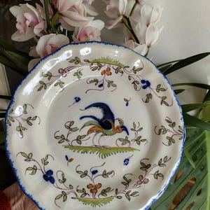 Peut inclure: Assiette en céramique blanche avec un bord festonné et un bord bleu. L'assiette présente un motif peint à la main d'un oiseau bleu et orange, entouré de feuilles vertes et d'éléments floraux. Une pièce décorative.