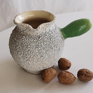 May include: A textured, ceramic pitcher with a green handle and a cream-colored rim. The pitcher is accompanied by four walnuts. The pitcher has a unique, handmade appearance, suggesting it is a decorative item.