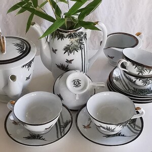 Peut inclure: Service à thé en porcelaine blanche orné d'un motif de bambous et d'oiseaux. L'ensemble comprend une théière, un sucrier, une crémière, ainsi que des tasses et soucoupes. Les bords et les anses sont dorés.