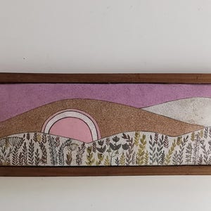 May include: A rectangular framed artwork featuring a landscape scene. The artwork depicts a pink sun setting behind brown and white hills, with a lavender sky. The foreground shows a field of stylized plants in shades of green and gray.