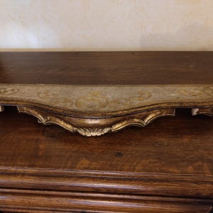 Peut inclure: Étagère décorative de style antique avec un bord avant incurvé et des détails dorés ornés. L'étagère est peinte dans une palette de couleurs or et grise, avec un motif décoratif sur la surface supérieure. Fabriquée en bois.