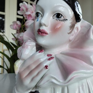 Peut inclure: Figurine de Pierrot en céramique avec un bonnet noir, un visage blanc et des joues roses. La figurine a une larme, des lèvres rouges et du vernis à ongles rouge. Elle porte un col rose et blanc à volants et tient une sphère noire.