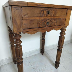 May include: A wooden side table featuring a rectangular top and two stacked drawers, each equipped with a central circular keyhole. The table is supported by four turned legs with vertical fluting details and a scalloped apron beneath the drawers.
