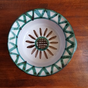 May include: A white ceramic bowl with a green and black geometric pattern around the rim. The bowl has a brown flower design with a small, square, metal strainer in the centre.