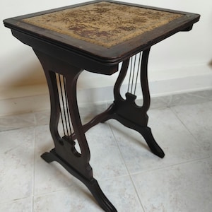 Peut inclure: Une table d'appoint en bois brun foncé dotée d'un plateau carré avec une incrustation beige texturée et usée. La table est soutenue par deux pieds en forme de lyre de chaque côté, reliés par de fines tiges métalliques verticales.