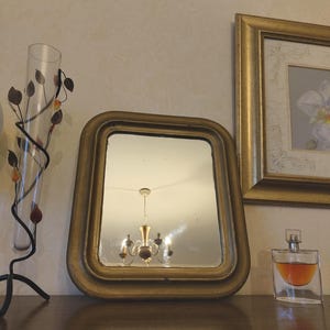 Peut inclure: Un miroir rectangulaire avec un cadre doré épais et arrondi est placé entre un vase en verre haut et fin sur un support en métal noir en forme de vigne avec des feuilles, et un flacon de parfum en verre contenant un liquide ambré.