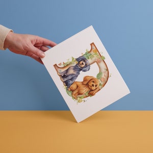May include: A white card with a watercolor illustration of two dogs sitting next to each other. The dogs are a blue and black spaniel and a brown and white spaniel. The dogs are sitting in front of a large letter D made of flowers.
