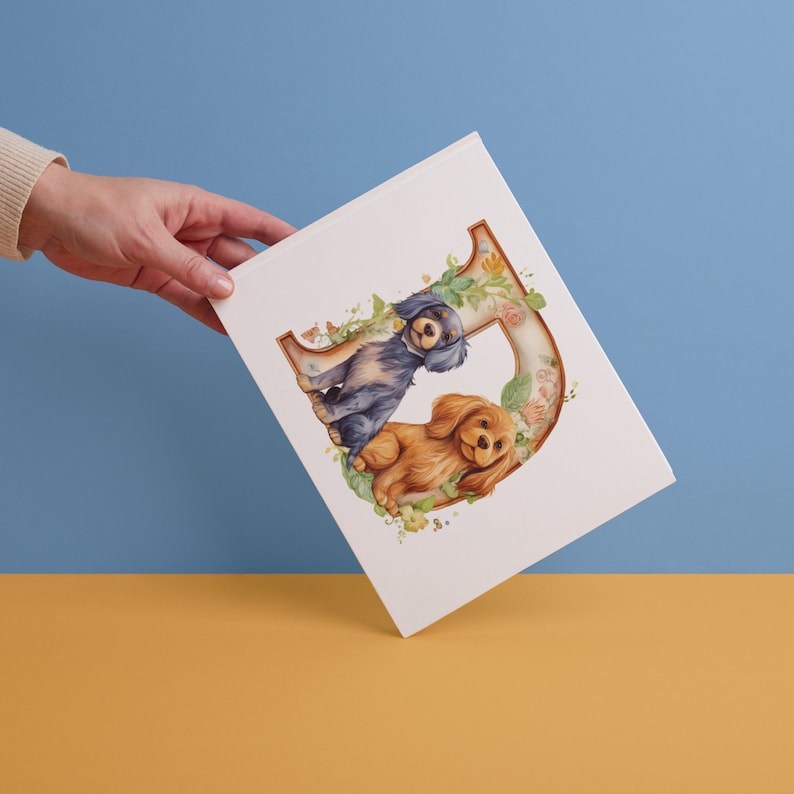 May include: A white card with a watercolor illustration of two dogs sitting next to each other. The dogs are a blue and black spaniel and a brown and white spaniel. The dogs are sitting in front of a large letter D made of flowers.