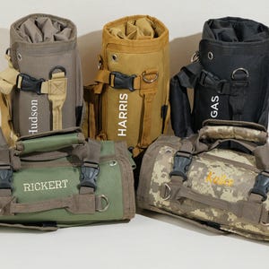 May include: Five personalized green, brown, and black canvas travel bags with adjustable straps and buckles. The bags are rolled up and have names embroidered on them: Rickert, Hudson, Harris, Gas, and Kyler.