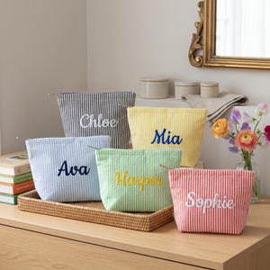 May include: Five striped cosmetic bags in navy, yellow, light blue, green, and red. Each bag is embroidered with a name: Chloe, Mia, Ava, Harper, and Sophie. The bags have a striped pattern and a zip closure.