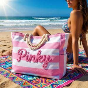 Op de afbeelding: Een grote roze en witte gestreepte draagtas met het woord "Pinky" in roze script. De tas heeft touwhandvatten en ligt op een kleurrijke strandhanddoek. De achtergrond toont de oceaan en een zonnige hemel.