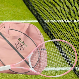 May include: A pink tennis bag with a tennis racket and the name "Robert" embroidered on it. The bag has a gray zipper and a gray strap. The racket is white with a pink handle.