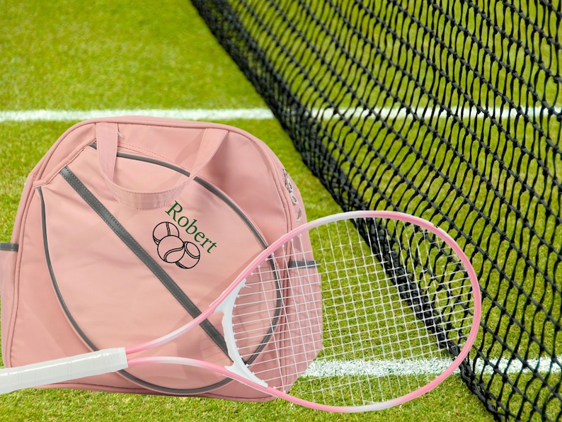May include: A pink tennis bag with a tennis racket and the name "Robert" embroidered on it. The bag has a gray zipper and a gray strap. The racket is white with a pink handle.