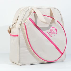 May include: A white tennis bag with a pink trim and a pink circle on the front with the text "The Bella Tennis Club" and a tennis racket graphic.