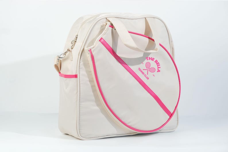 May include: A white tennis bag with a pink trim and a pink circle on the front with the text "The Bella Tennis Club" and a tennis racket graphic.