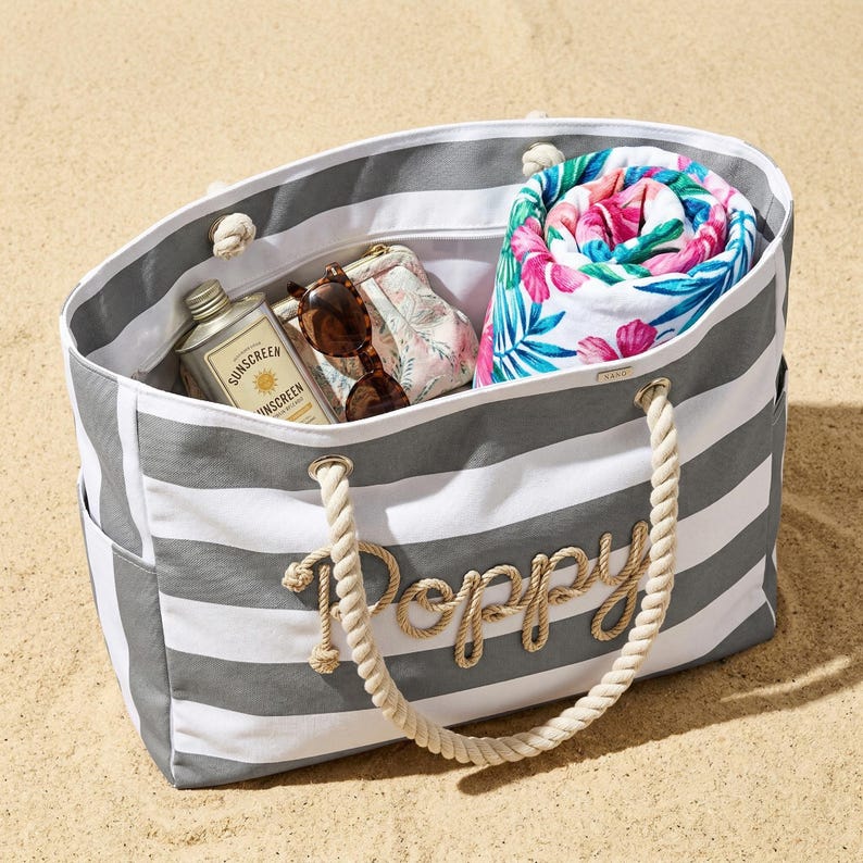 Peut inclure: Un sac de plage &agrave; rayures grises et blanches avec le nom "Poppy" en lettres de corde. Le sac contient une serviette de plage fleurie enroul&eacute;e, des lunettes de soleil, une petite pochette &agrave; fleurs et une bouteille de cr&egrave;me solaire.