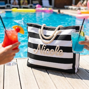 Peut inclure: Un sac fourre-tout &agrave; rayures noires et blanches avec le nom "Nicole" brod&eacute; en lettres de style corde. Le sac a des poign&eacute;es en corde et se trouve pr&egrave;s d'une piscine avec deux cocktails.