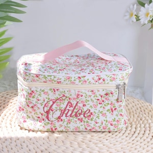 Könnte beinhalten: Eine Kosmetiktasche mit floralem Muster, rosa Griff und Reißverschluss. Die Tasche ist weiß mit einem Muster aus kleinen rosa und roten Blumen und grünen Blättern. Der Name "Chloe" ist in rosa Schrift auf der Vorderseite gestickt.