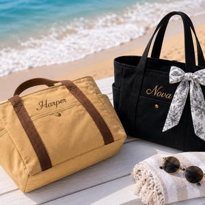 Op de afbeelding: Een beige canvas tas met bruine hengsels en de naam Harper in bruin geborduurd, en een zwarte canvas tas met de naam Nova in goud geborduurd, voorzien van een wit-zwarte strik met bloemenpatroon en een ronde zonnebril.