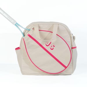 May include: A beige canvas tennis bag with a pink trim and a pink tennis racket outline. The bag has a zippered pocket and a handle. The initials "JS" are embroidered on the front of the bag.