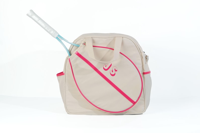May include: A beige canvas tennis bag with a pink trim and a pink tennis racket outline. The bag has a zippered pocket and a handle. The initials "JS" are embroidered on the front of the bag.