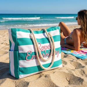 Può includere: Una grande borsa tote con strisce orizzontali color verde acqua e bianco, due spessi manici in corda beige e la scritta "Rope Embroidery" ricamata con filo rosa sul davanti. La borsa presenta tasche laterali ed è appoggiata sulla sabbia.