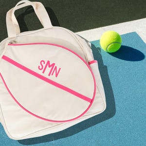 May include: A white tennis bag with a pink trim and the initials "SMN" in pink lettering. The bag is shaped like a tennis racket and is lying on a blue tennis court with a yellow tennis ball.
